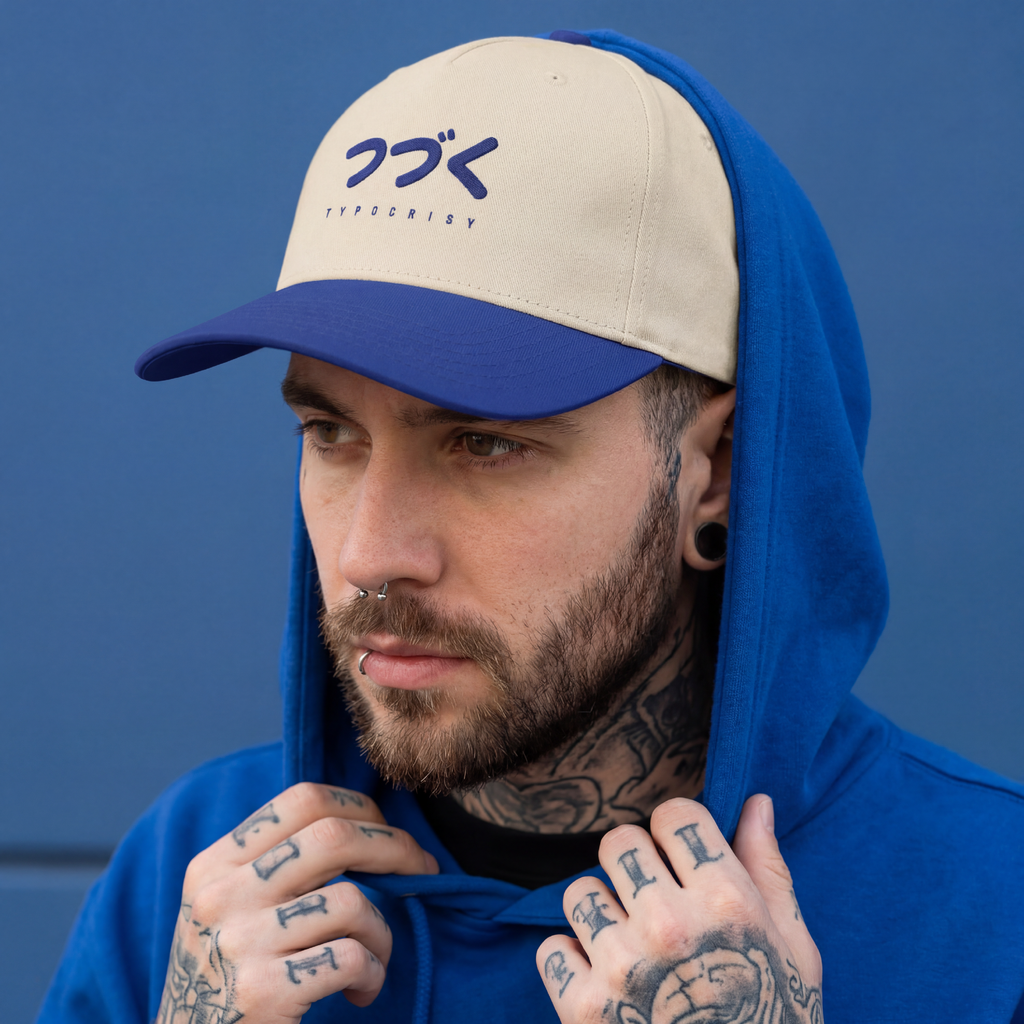 Man wearing a blue and beige cap with a logo, blue hoodie, and tattoos on his arms against a blue background.