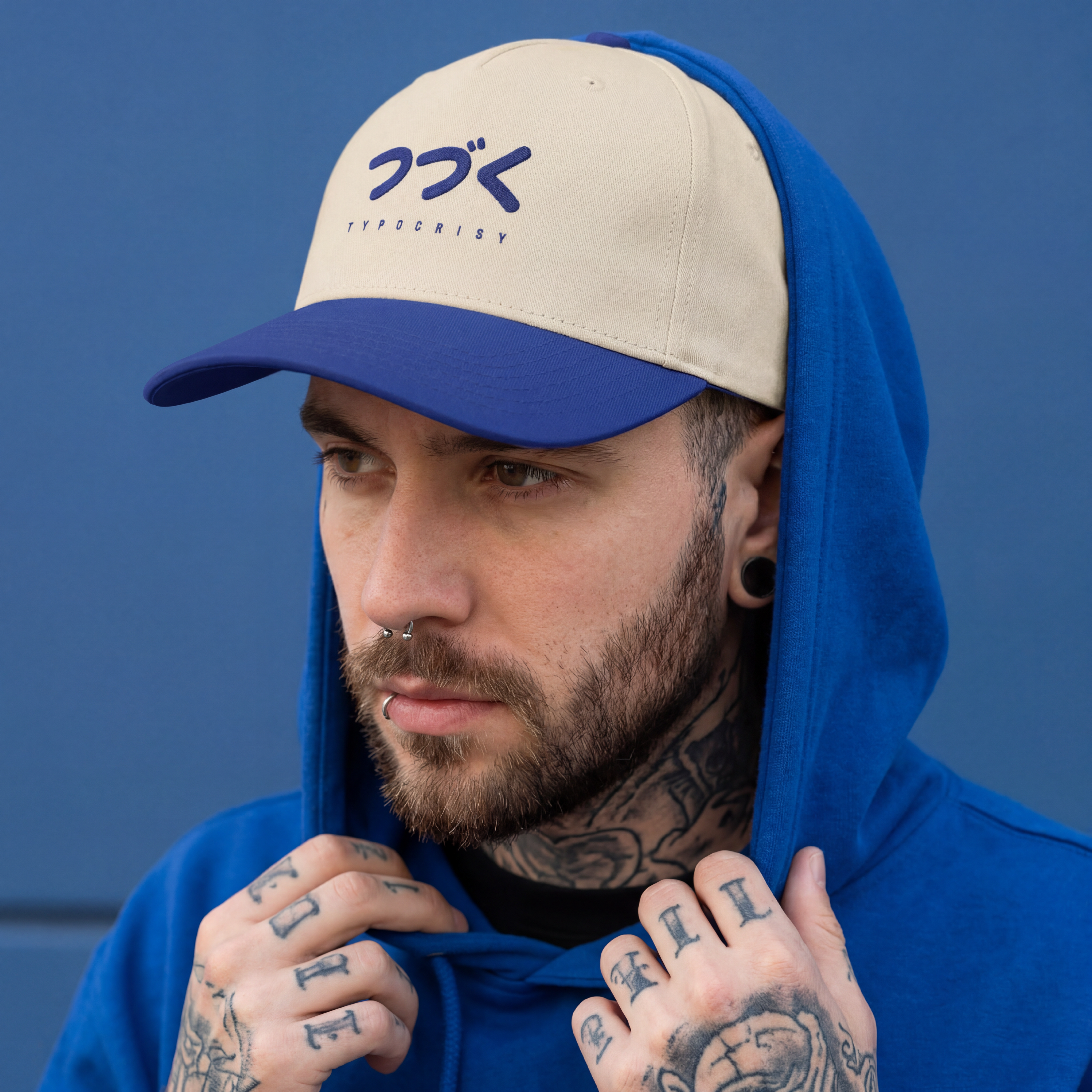 Man wearing a blue and beige cap with a logo, blue hoodie, and tattoos on his arms against a blue background.