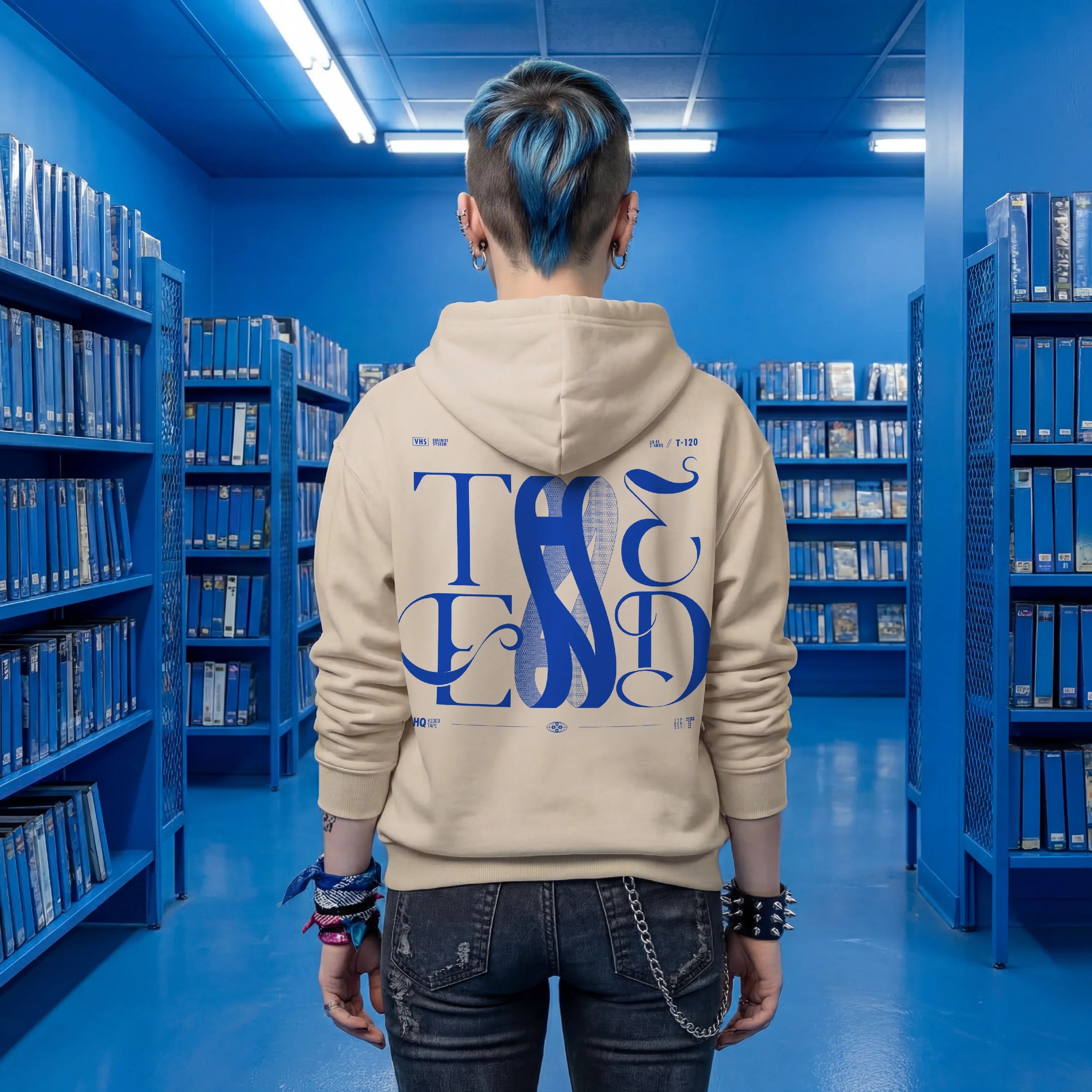 Beige oversized hoodie with bold blue THE END baroque typography graphic on the back, worn by a person with blue hair standing in a VHS archive aisle — streetwear by TYPOCRISY