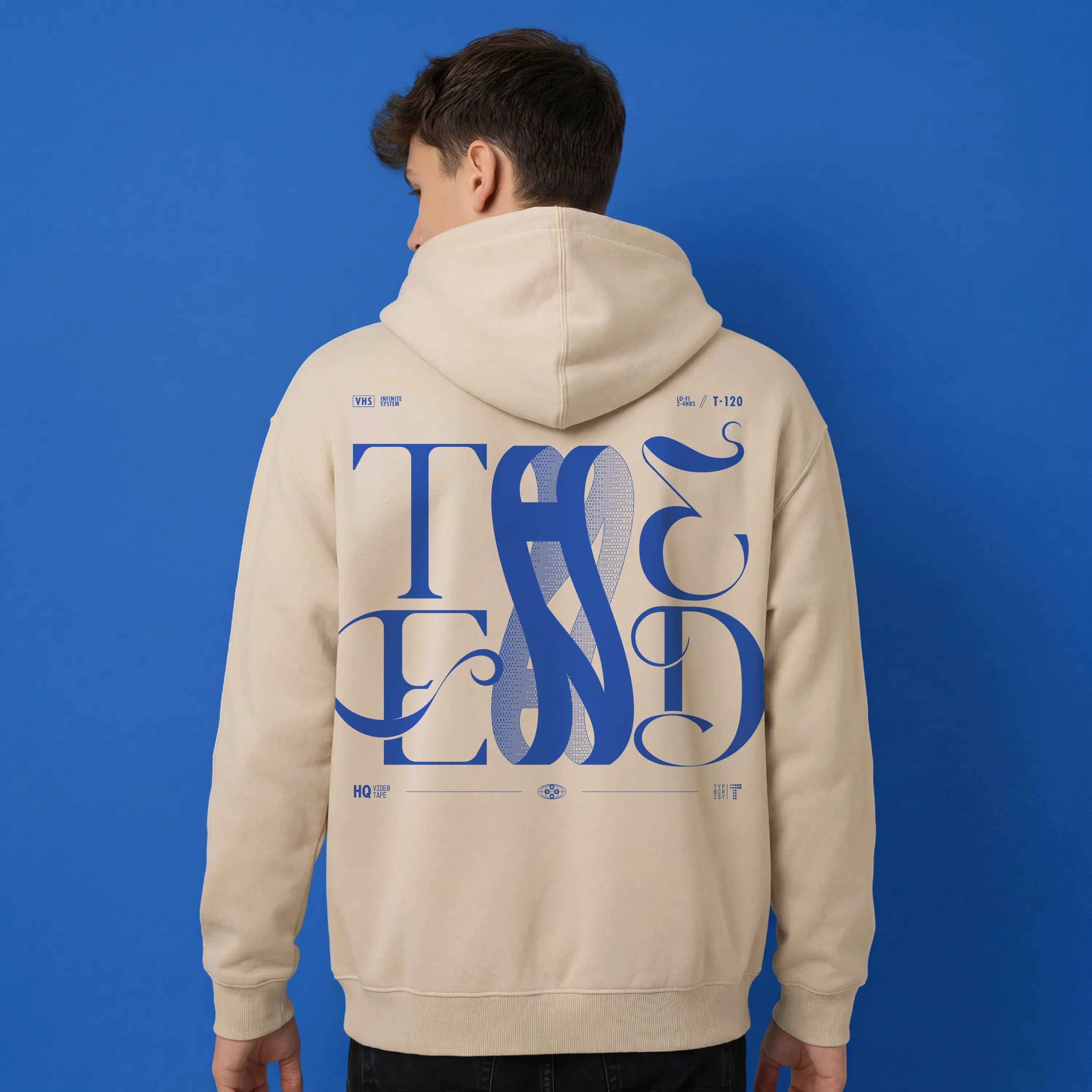 Back view of beige oversized hoodie with large blue THE END interlocking baroque typography graphic, worn by a man against a cobalt blue background — THE END Graphic Hoodie by