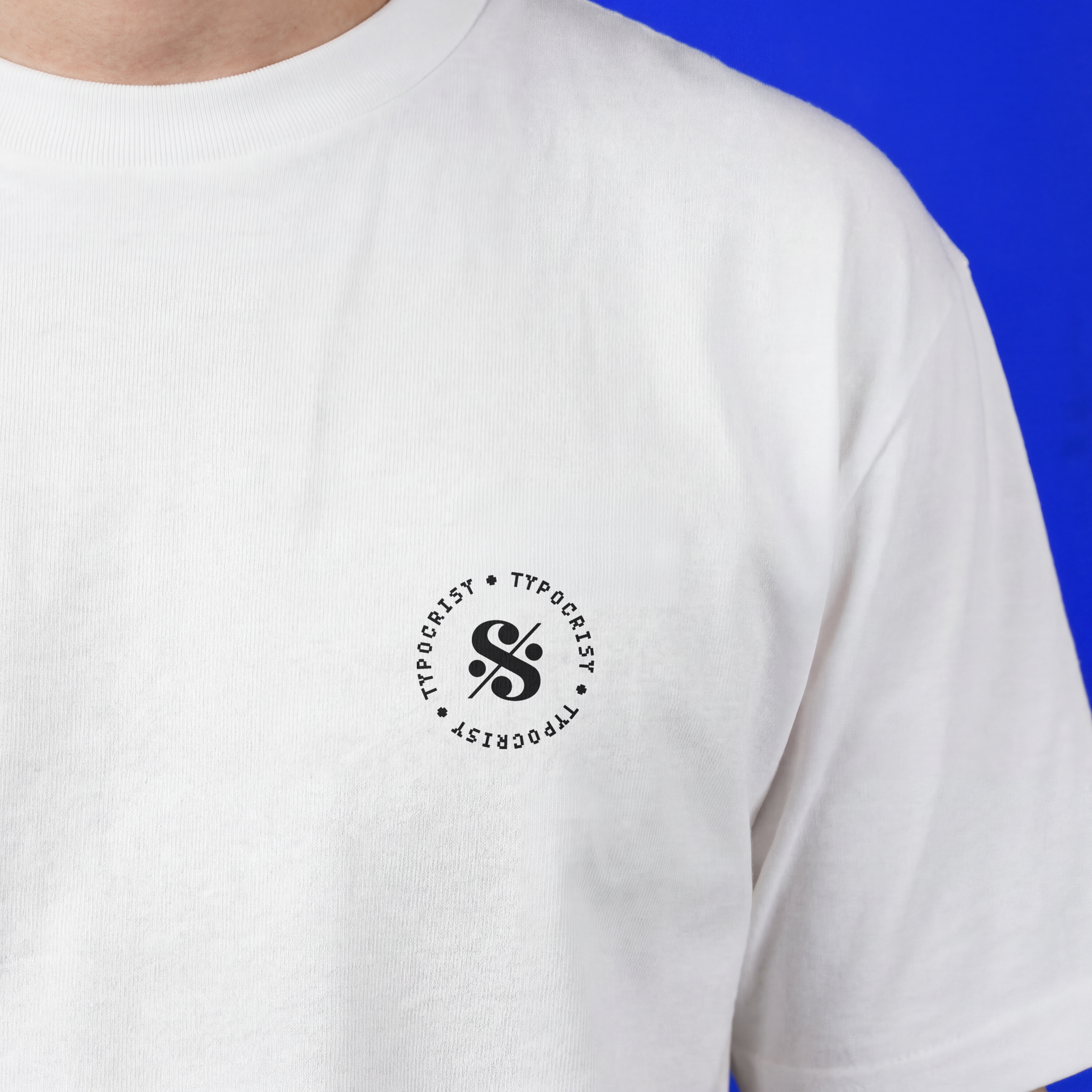 Close-up of white Typocrisy SIL3NC3 oversized t-shirt front chest, circular Typocrisy logo badge with stylized S mark, blue background