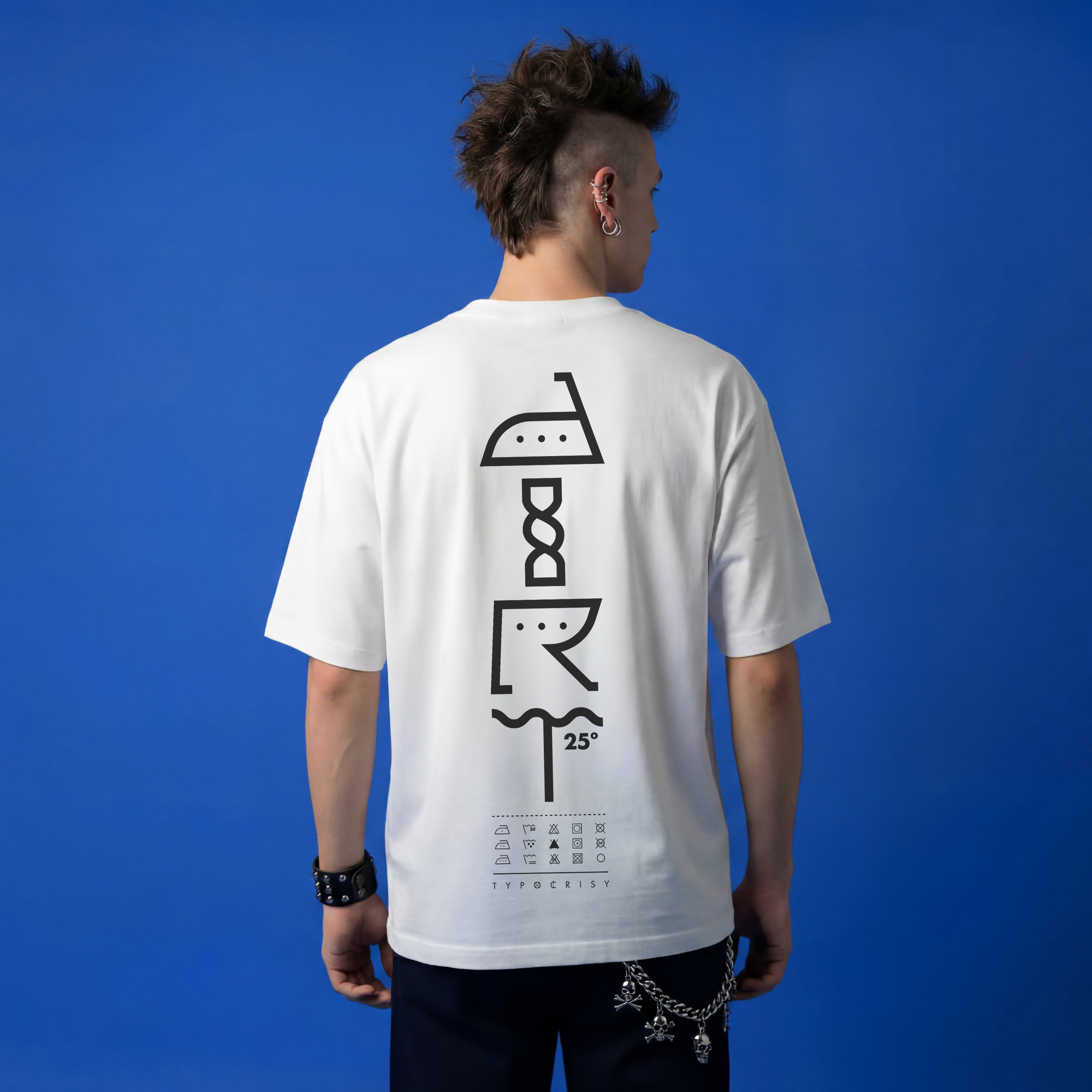Man with mohawk wearing white Typocrisy DIRT 25 oversized t-shirt, back graphic spelling DIRT in laundry care symbols with 25° wash temp and TYPOCRISY wordmark, blue studio background