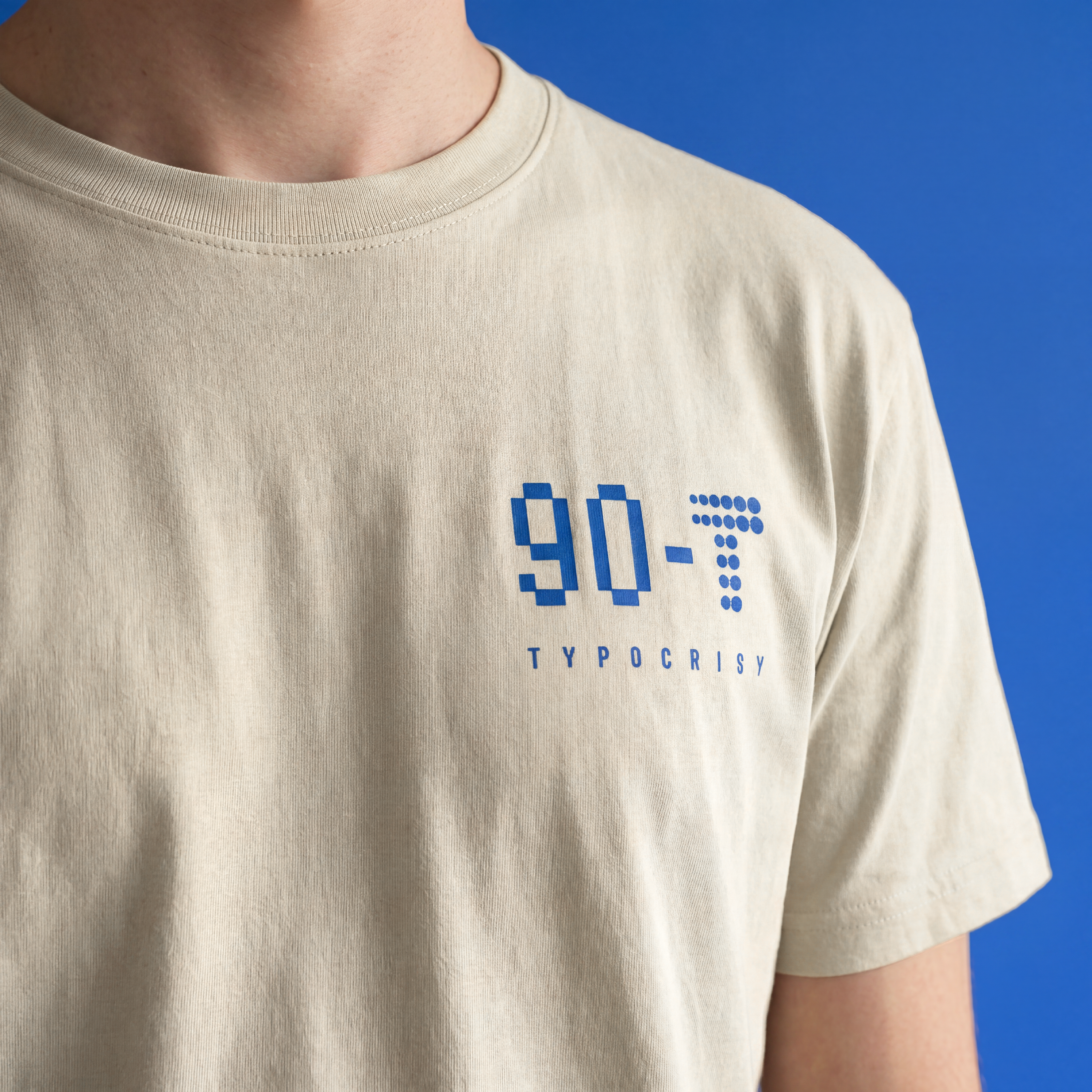 A close-up of a person wearing a light beige crewneck t-shirt with a blue pixelated '90-T' and 'TYPOCRISY' graphic on the chest, set against a solid blue background.