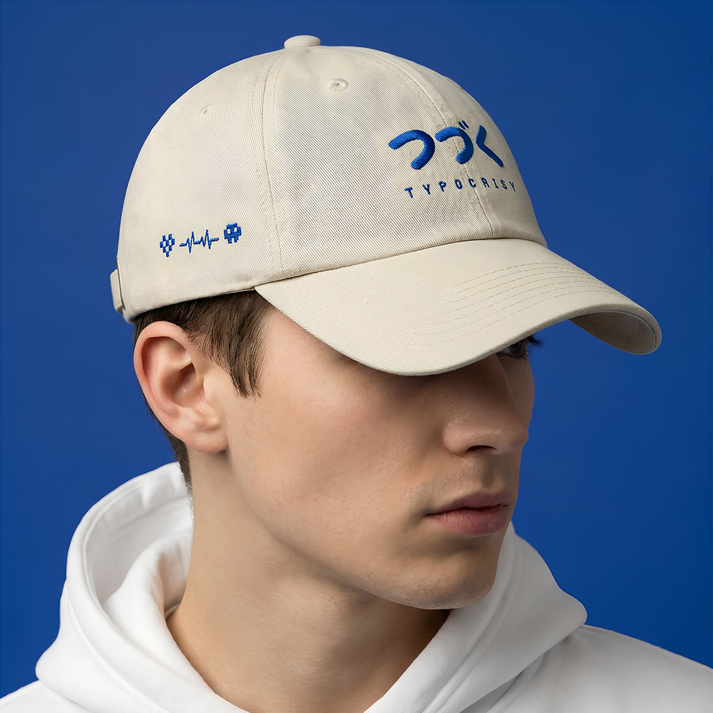 Person wearing a Typocrisy つづく To Be Continued Baseball beige cap with blue branding against a blue background