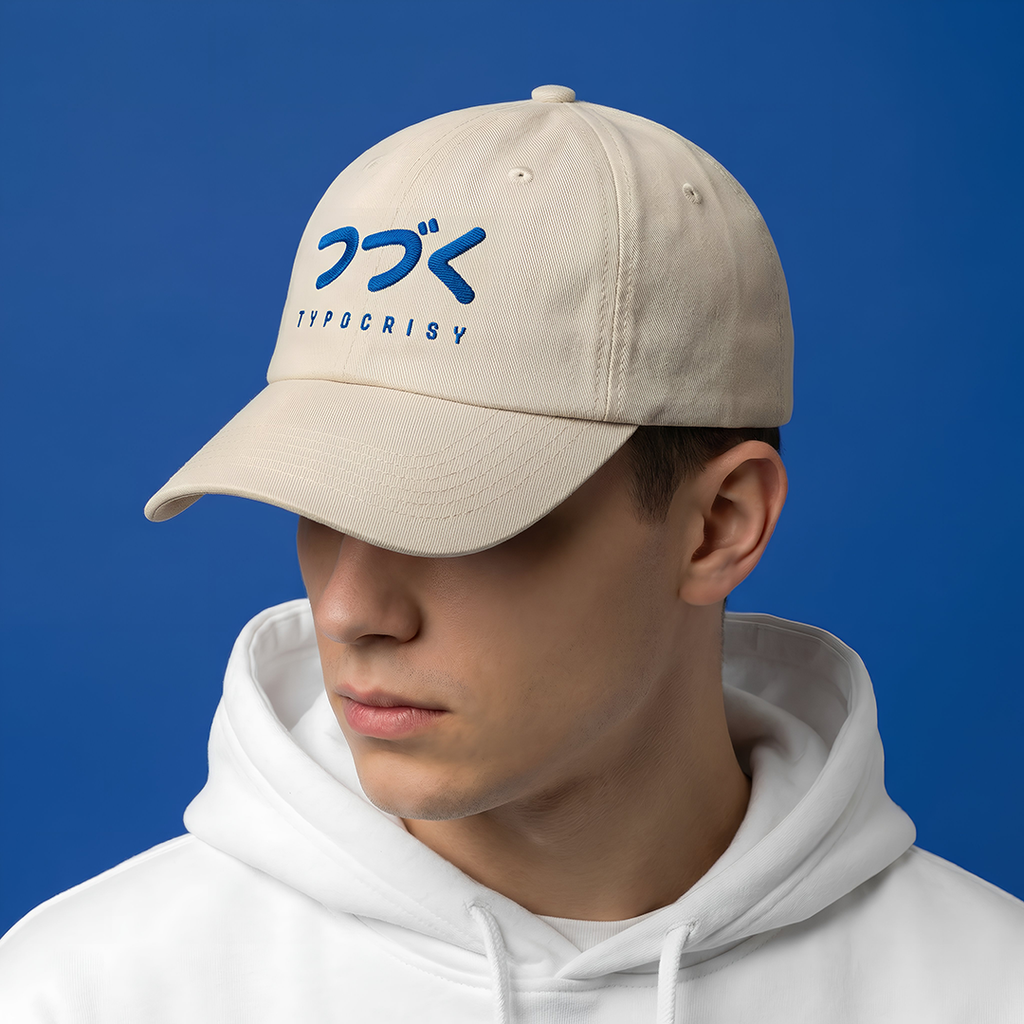 Person wearing a Typocrisy つづく To Be Continued Baseball beige cap with blue text on a blue background