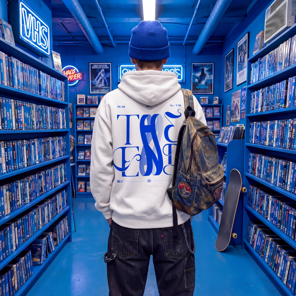 White oversized hoodie with bold blue THE END baroque typography graphic on the back, worn by a man carrying a camo backpack and skateboard in a neon-lit VHS store — retro streetwear by TYPOCRISY