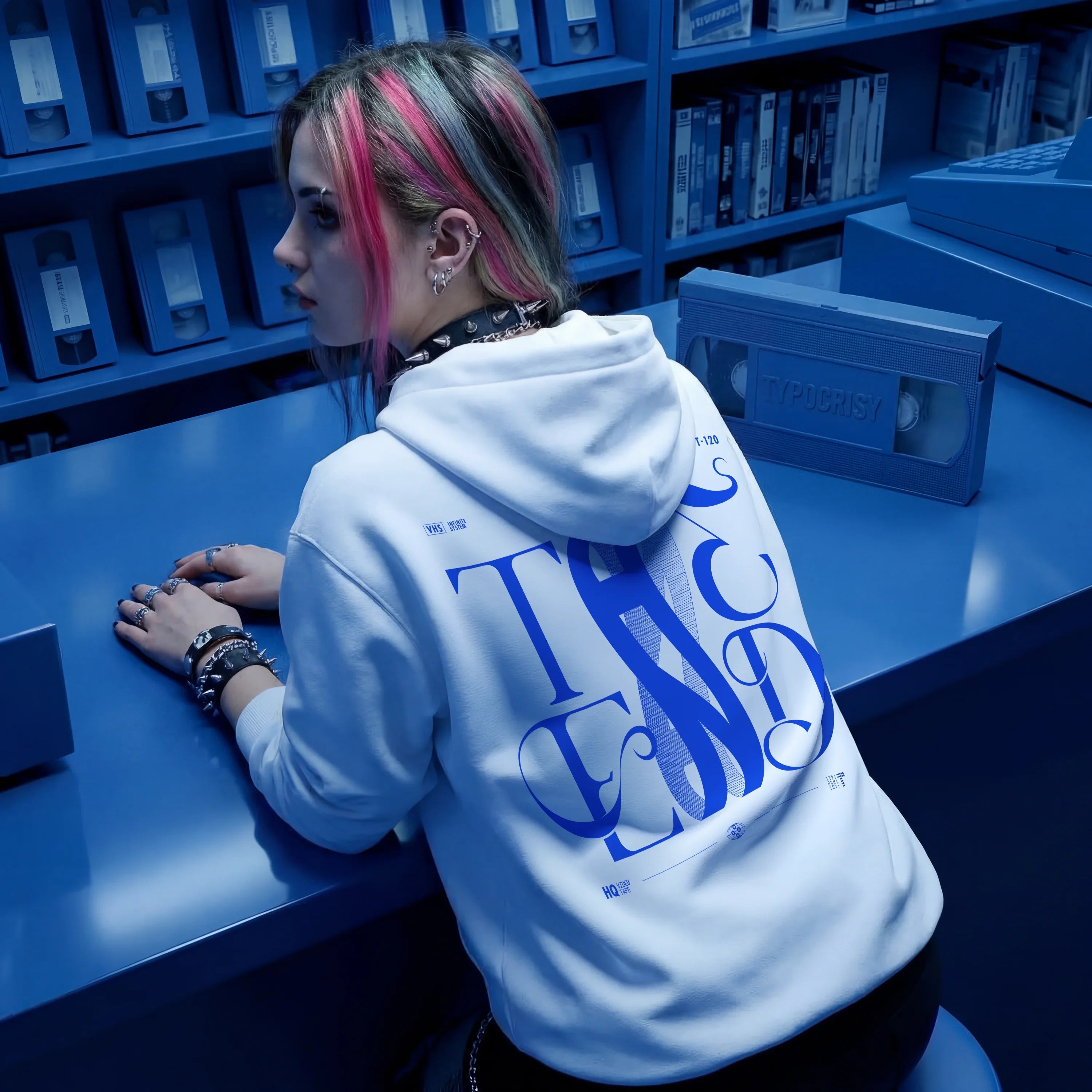 White oversized hoodie with bold blue THE END baroque typography graphic on the back, worn by a woman with pink-streaked hair in a blue-lit VHS video store — retro streetwear by TYPOCRISY
